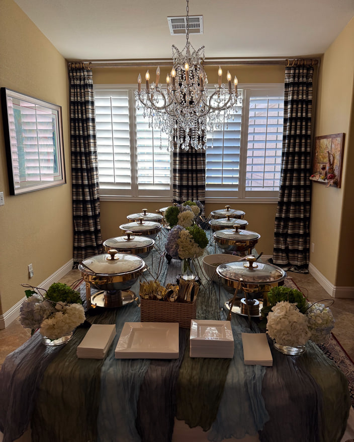 A long buffet table set with chafing dishes and greenery under a chandelier.