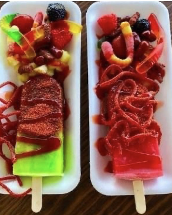 Two fruit paletas on white plates, drizzled with chamoy and chilli powder and piled with gummy sweets, strawberries and mango.
