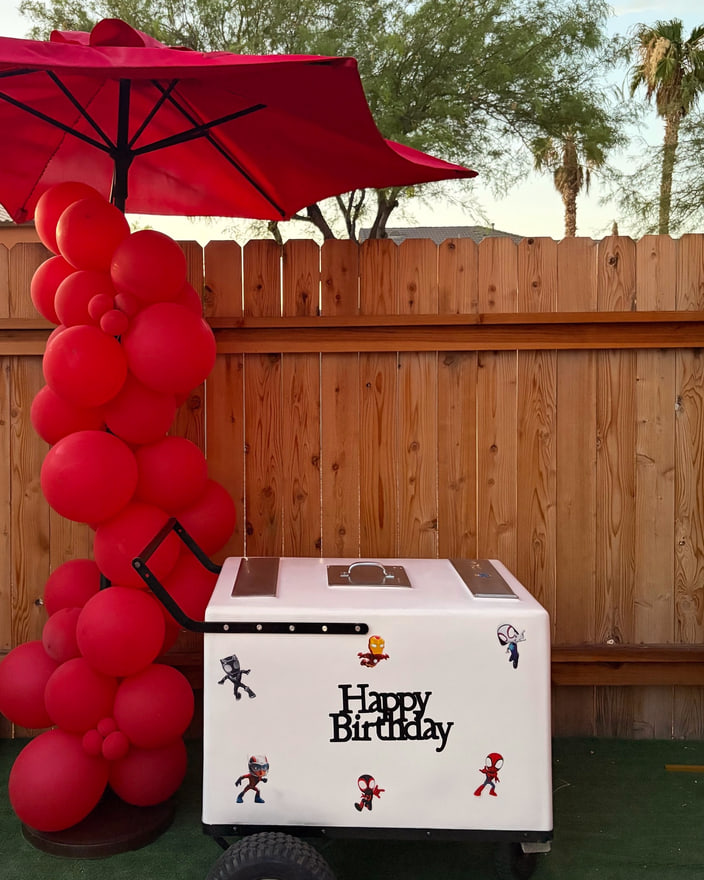 A paleta cart styled with red balloons and a red umbrella against a wooden fence.