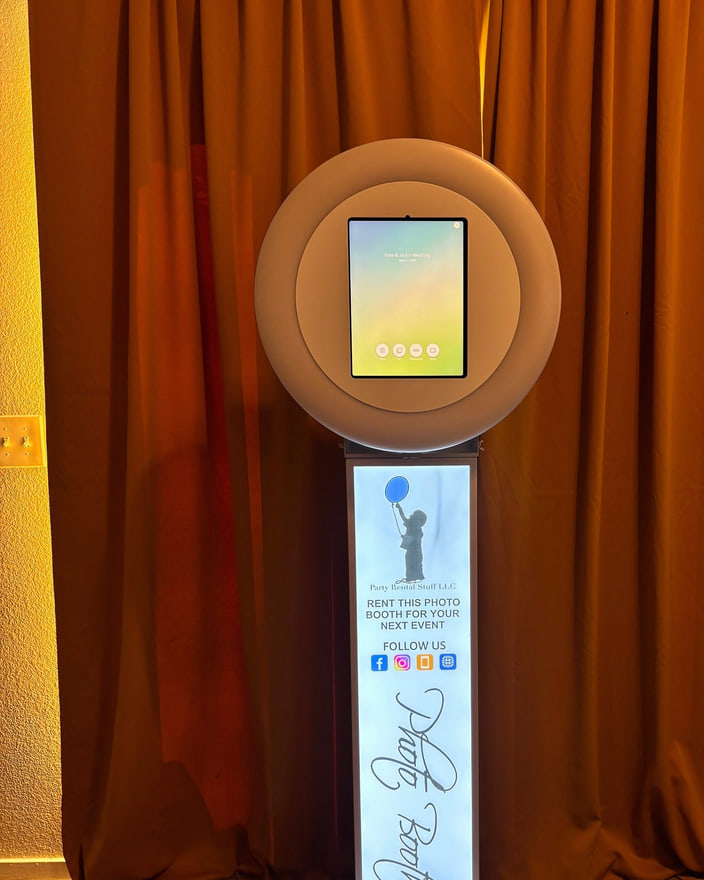 An open-air photo booth with a lit ring light and a tablet screen, standing against a gold drape.