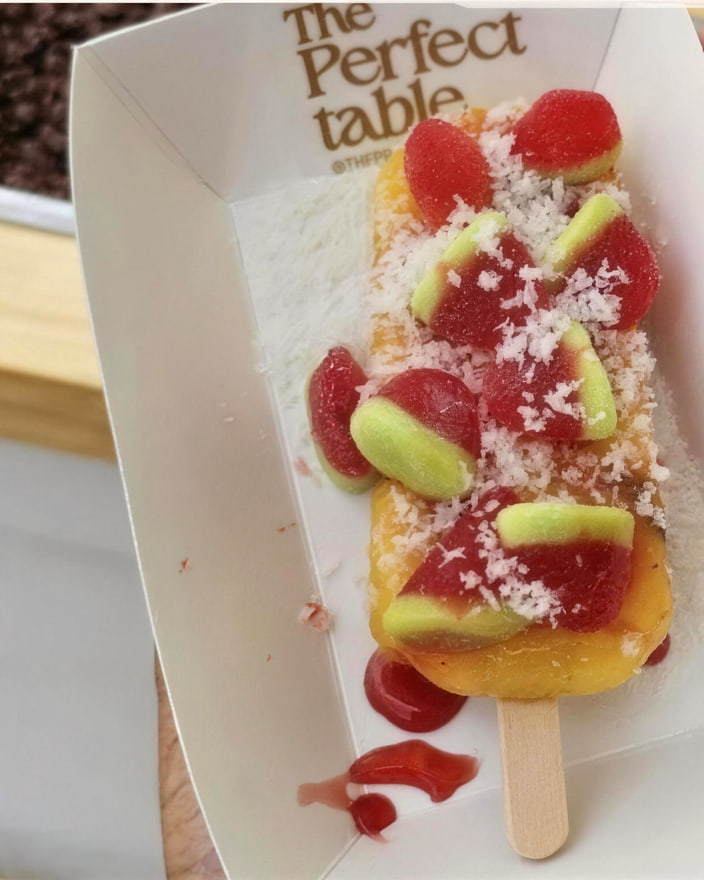 A mango paleta in a paper tray, covered with shredded coconut, chamoy and watermelon gummy sweets.